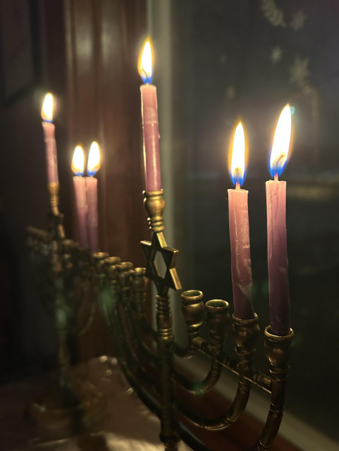 Two traditional brass chanukiyot photographed from an angle, with purple dipped candles lit for night two. They are in front of a window and surrounded by darkness. The brass glows faintly from the candles.