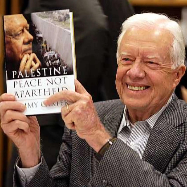 Jimmy Carter smiling as he holds up his book titled Palestine: Peace not apartheid.