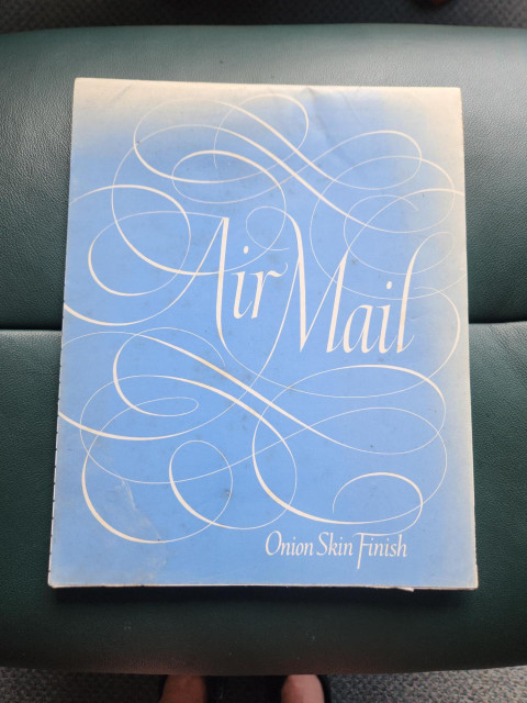 An ancient blue note pad bearing the words "Air Mail" in large cursive writing amid decorative swirls, and the words "Onion Skin Finish" in smaller cursive writing on the bottom right hand side. The cover of the notepad has been bleached by exposure to sunlight across the top and right edge.