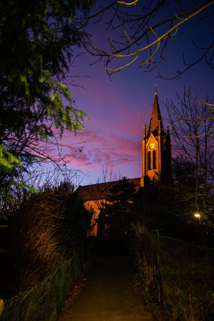 Das Foto zeigt den Turm unserer Dorfkirche, der von einem Strahler angeleuchtet wird. Im Hintergrund zeigt sich der rote Himmel des Sonnenaufgangs.
Rechts und links ragen Teile von Bäumen in das Bild hinein, die an dem schmalen Weg liegen, der hinauf zur Kirche führt. 