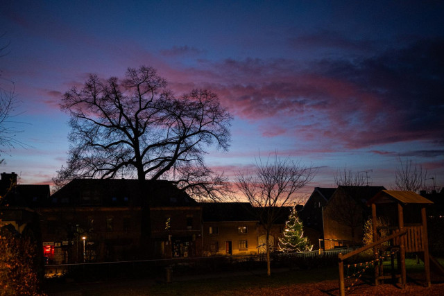 Das Foto zeigt einen Platz in unserem Dorf. Es steht ein Baum auf dem Platz und ein noch beleuchteter Weihnachtsbaum. Im Vordergrund rechts ist noch ein Häuschen eines Kinderspielplatzes zu erkennen.
Im Hintergrund Häuser und ein paar Strassenlaternen.
Der Himmel ist blau und Teile der Wolken sind schon rot durch den bevorstehenden Sonnenaufgangs.