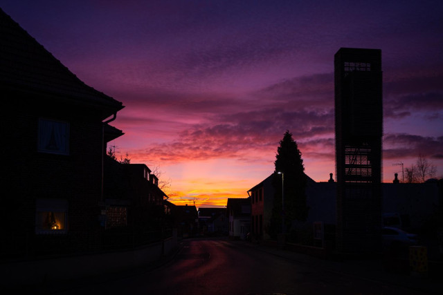 Silhouette von Häusern an unserer Hauptstrasse, mit dem Kirchturm der evangelischen Dorfgemeinde. 
Der Sonne geht langsam auf, so das der Himmel in Teilen schon rot ist. 