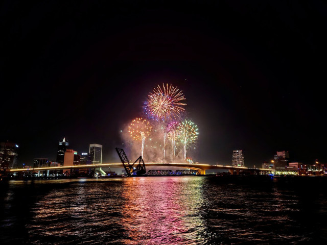 Burst of sparkly and colorful fireworks shooting up into the sky over a dark river at night with an illuminated city skyline as the background.