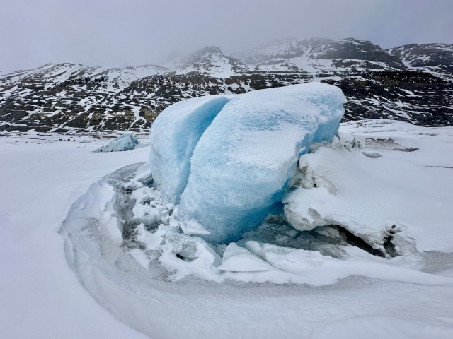 A cracked glacial blue ice formation is surrounded by a swollen frozen lake, with rocky mountains in the background shrouded in misty clouds. Remains of the Saskatchewan glacier.  