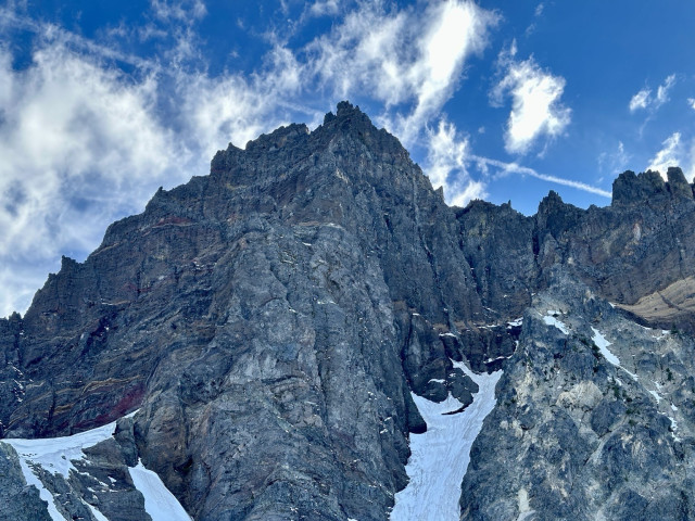 A rugged mountain landscape featuring steep rock faces and patches of snow, under a blue sky with scattered clouds. East face of Three Fingered Jack. 