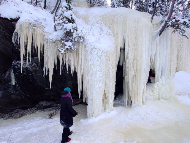 A figure in a black parka & blue hat stands before a wall of ice formed from water trickling over a rock face.