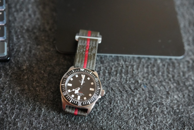 A black dial Tudor Pelagos FXD on its original green with red stripe velcro strap. It’s partially sitting on a black Magic Trackpad.