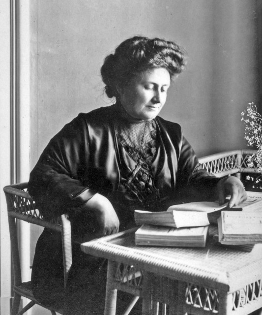 Posed photo of Maria Montessori at a small desk. She is typically dressed for a white middle-class European of the time.