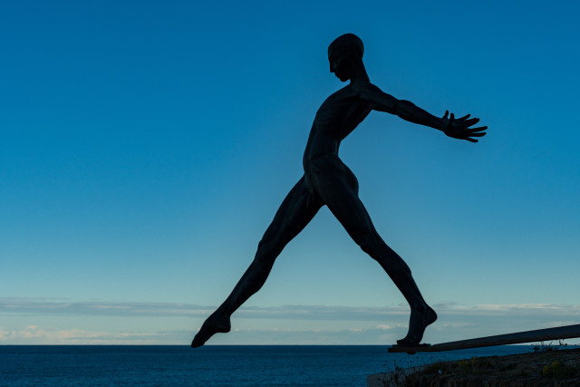 Ein Foto einer nackten männlichen Bronzeskulptur mit dem Namen ' le Défi' im französischen Antibes. Der Standort lässt den Blickwinkel so zu dass der Eindruck entsteht, die Skupltur würde mit den Zehen das Meer berühren. 