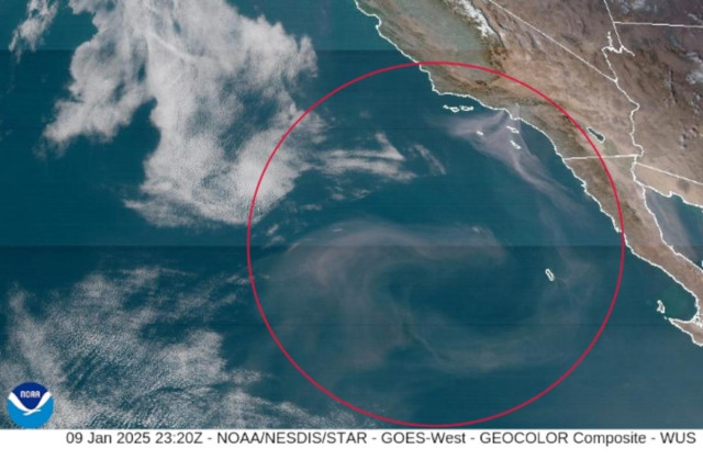 A color satellite photo showing southern California in the upper right and the Pacific ocean in the rest of the frame. There are a few clouds in the upper left. A large red circle has been added to show a large swirling plume of smoke from the catastrophic fires in the Los Angeles region. The smoke fans out from two prominent location in the LA region and then swirls south toward the bottom of the frame before swirling into a clockwise spiral around the bottom center right.