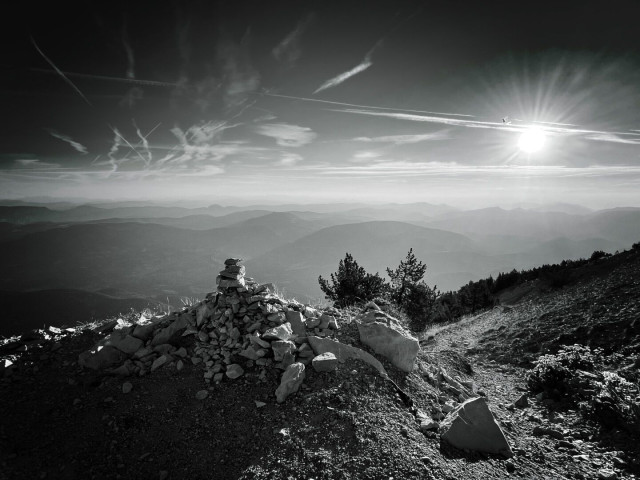 A black-and-white landscape featuring a rocky outcrop and a pile of stones in the foreground, with a panoramic view of rolling mountains under a partly cloudy sky. The sun is low, casting rays across the scene.