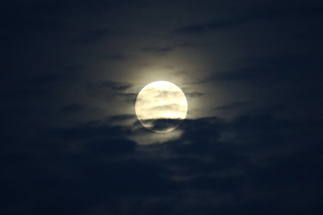 Vollmond hinter Wolken. Der kreisrunde, hell leuchtende Mond wird zum Teil von vorbeiziehenden Wolken verdeckt. Um ihn herum ist der Himmel gelb illuminiert.