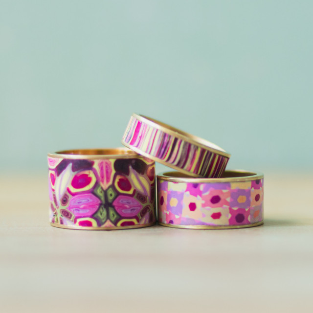 Three brass channel rings, with different coordinating polymer clay designs featuring pink, purple and yellow, inlaid into the channel.