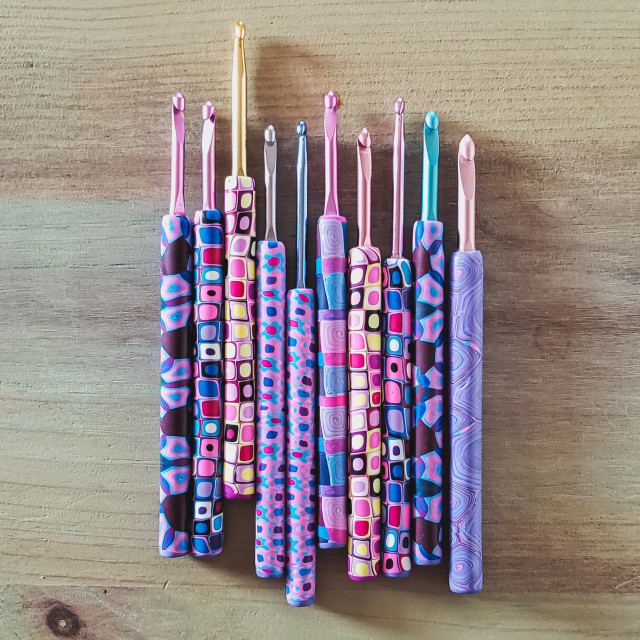 A selection of metal crochet hooks with colorful, decorative ergonomic polymer clay handles, in designs featuring pinks and purples.