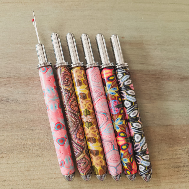 A selection of sturdy chrome seam rippers, one open and five closed, featuring colorful polymer clay handles in pinks, golds, and other colors.