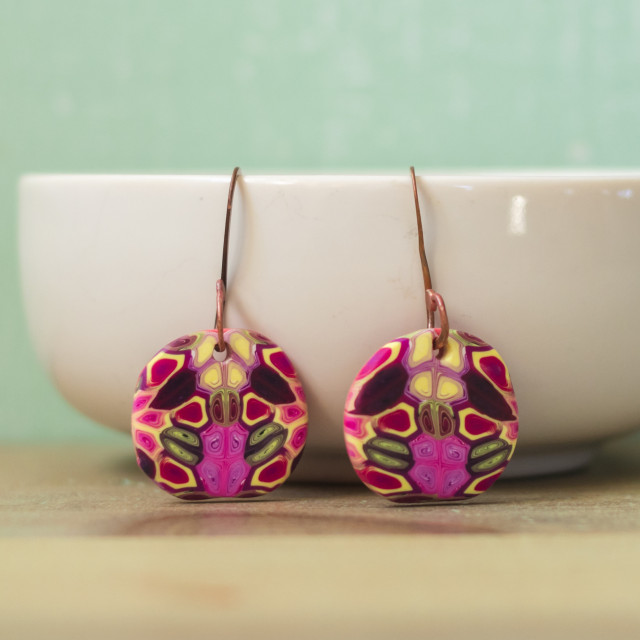 A pair of earrings comprised of handmade copper earring wires, and round polymer clay tiles with a kaleidoscopic design in pink, yellow, and purple.