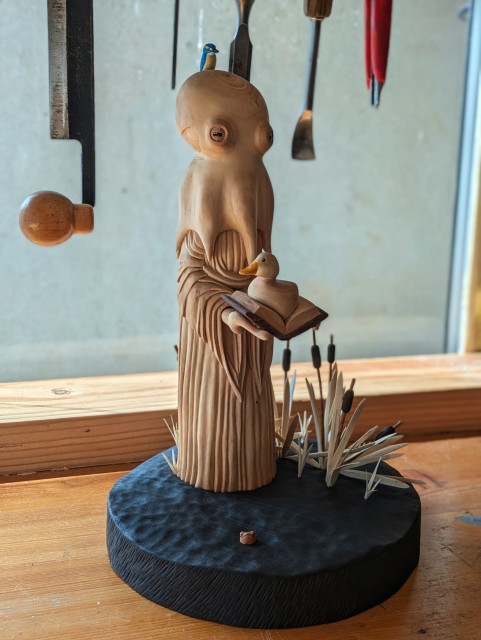 Woodcarved humanoid octopus wearing a dress, holding a book with a duck on it. It has a kingfisher on it's head. The base is a dark pond, there is a tiny head of an unhappy frog and some bulrushes