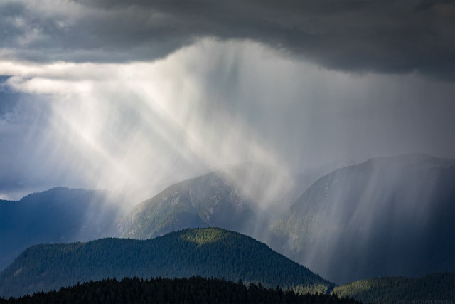 A downpour of rain over mountains with rays of sunlight shining in from the left.