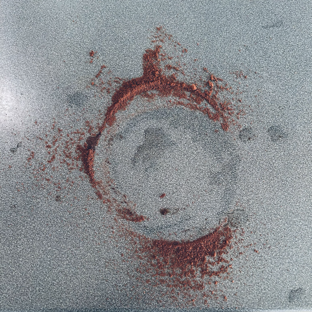 A square photo in colour shows a speckled-grey countertop in a well-lit kitchen. On the countertop's surface is spilled espresso, ground finely as suitable for a stovetop moka-pot. The espresso-grounds' shape has been unaltered from where it fell naturally. The espresso grounds are splayed in a sort of a round shape, feathering at the edges, as was created when the coffee overflowed out of the moka-pot's coffee holder & onto the countertop. The result is an open-circle shape, almost like a calligraphic sphere as when made with ink in what is called Enso practice. Despite any alteration of plans that the spilled coffee may have caused, the moment of the photo is completely content & cheery, with a happy feeling of much-needed pause & relaxation.