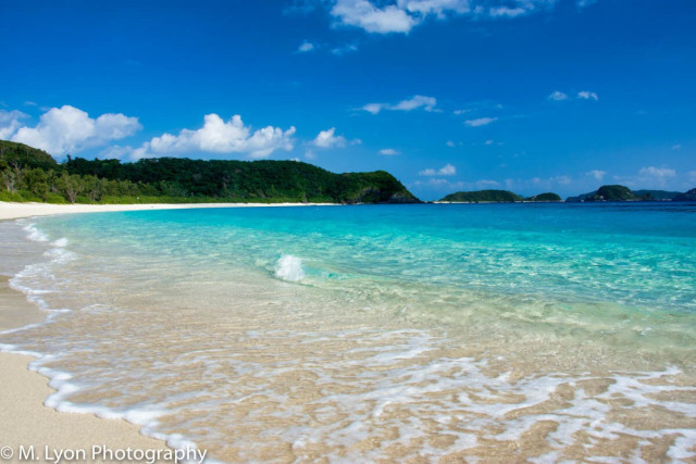 A serene beach scene with clear turquoise water lapping gently against a sandy shore. Lush green hills are in the background under a bright blue sky with a few scattered clouds.