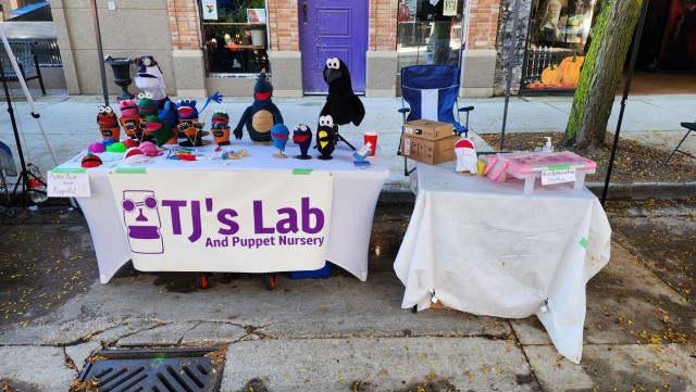 A booth at a vendor market. There's a large table with a banner reading "TJ's Lab and Puppet Nursery". The table is covered in small puppets, blank puppet bodies, and accessories for puppets. Behind the table are three larger puppets: A crow, a green dragon, and TJ himself (sporting his steampunk goggles).

A smaller table to the side labeled "customization station" has a tacklebox full of craft supplies.

The booth is set up outside on the edge of a street downtown. The shops in the background are decorated for Halloween, and there's leaves everywhere.