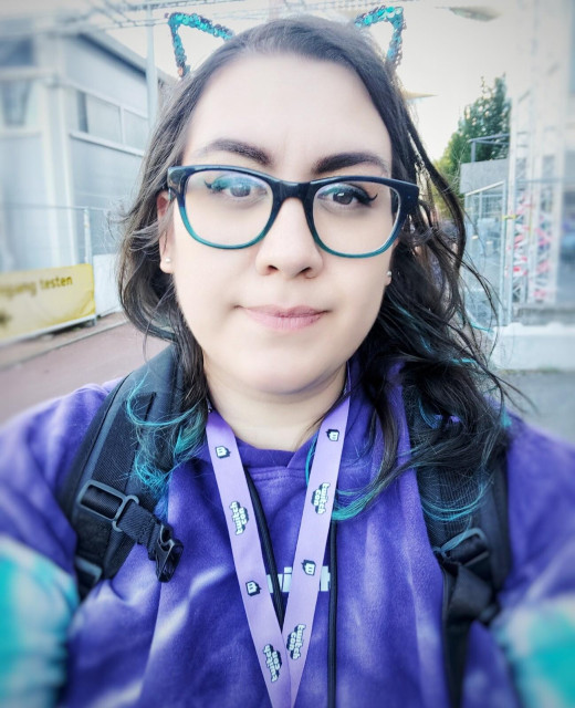 Rosie is a plus size Dutch Indonesian woman with a light to medium skin tone, brown eyes and dark brown hair. In this photo she has dark brown hair with turquoise ends and she has black eyeliner on. She is wearing black frame glasses, and purple and turquoise Twitch hoodie. Around her neck is a Twitchcon keycord / lanyard, and she is wearing a black backpack. She is looking into the camera and smiling faintly. On her head is a turquoise sequin cat ear tiara. She is outside, and behind her are some buildings and fences.