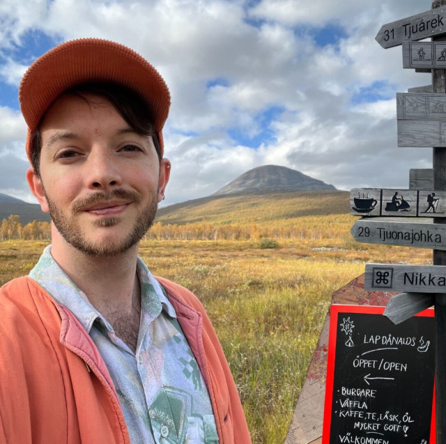 This is a selfie of me, a thirty two year old old with dark hair and a small beard. I am smiling whilst wearing an orange cap and a matching orange jacket. I am stood in front of an Arctic mountain in the late summer. There is a sign beside me advertising Lappdånals (a joke referring to Sámi McDonald’s), there is also a wooden sign post pointing to various Norrland places.