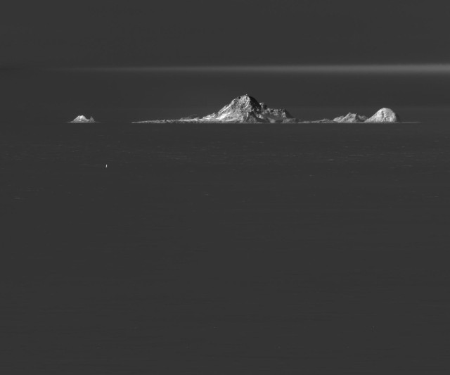 Greyscale image of Southest Farallon Island and Hurst Shoal, some significant rocks (mountains?) peaking out of the Pacific Ocean in the City of San Francisco. A layer of clouds frames the top of the image in he distance. A navigation buoy sits much further in the foreground. 