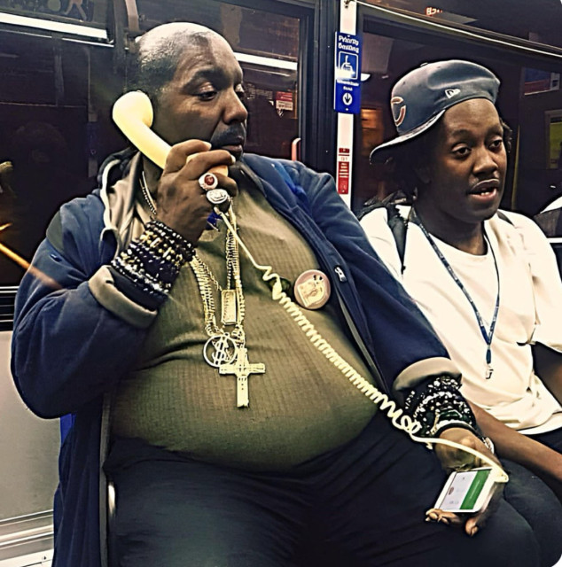 A man sitting in what appears to be a bus or a subway, holding an older style iPhone, but attached to the headphone jack is an old style telephone receiver held to his ear.  