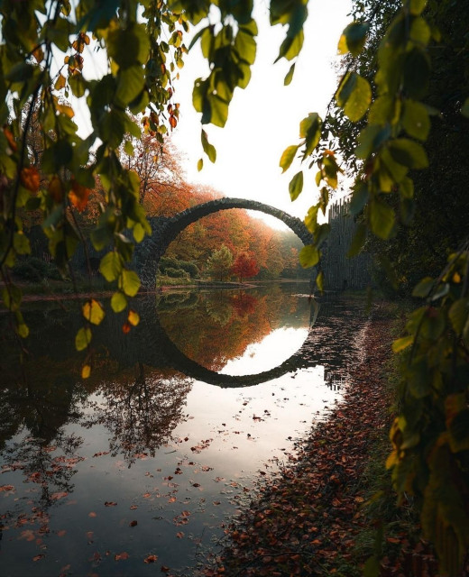 Photography.  A color photo of an old arched bridge over a lake in an autumnal landscape. The bridge is bent upwards and, together with the reflection in the water, forms a circle. The sun's rays falling from above change the lighting conditions in this circle. All around are autumnal deciduous trees on the bank. A beautiful nature shot and the photo has a wonderful atmosphere
Info: The 19.80-meter cantilever arch bridge was built by a landowner beginning in 1866 as part of a garden ensemble and is also known as the Devil's Bridge. The bridge was featured in the Hollywood film "Matrix Resurrections" and the 2017 German film "Der Zauberlehrling".
