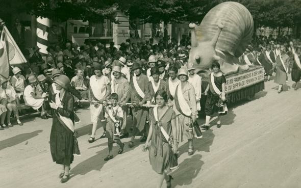 1920s suffragists marching through a street. They are pulling a parade float that carries a giant paper mache snail