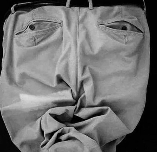 Photography. A black and white photo of the back of a pair of light-colored, wrinkled suit trousers. The trousers also have pockets at the back with small buttons as fasteners. The trousers are spread out, the many folds and the buttons (eyes) make the whole thing look like a wrinkled face with a long nose and mouth. A very nice photo as an example of pareidolia, a phenomenon in which people see faces or other patterns in normal everyday objects.