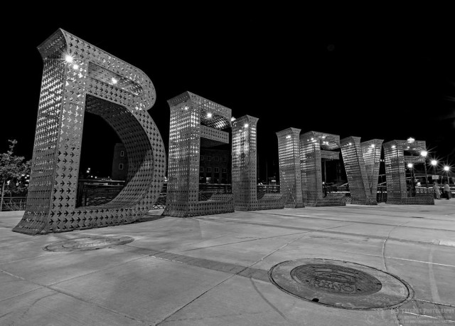 A black and white photo of a large metal sign. It is made up of individual letters. The metal has what appears to be small birds of planes cut into it. There a small lights built into the to part. The letters spell out the word, "Believe." The letters are arranged in a semi-circle. They are about ten feet tall. A manhole cover is in the right foreground. It says, "Reno," on it. 