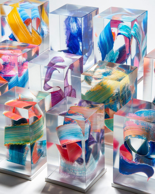 a series of sculptures, photographed in three-quarter view, made from cast clear resin in a cube form with three-dimensional multicolored brushstrokes inside, set against a white background