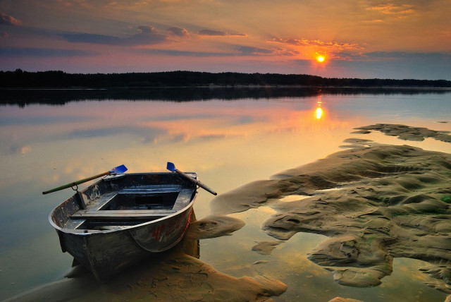 a boat at the sandy shore of a calm lake. Sunrise sun is lighting the clouds, painting them pink and reflecting in the still water.