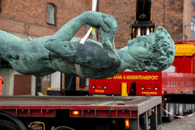 Greenish bronze sculpture of David is being moved by a crane in a horizontal position.