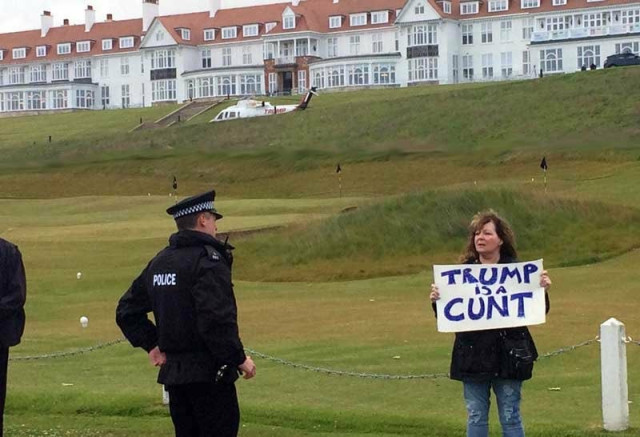 A photo taken in front of the Scottish Turnberry golf course, that Trump bought in 2014.  A middle aged woman is standing in front of the very grand clubhouse.  She is holding a sign in protest to Trump.  It reads; 'Trump is a cunt'.  A policeman is standing in front of her.  He isn't doing much (he probably agrees). He looks like he feels he should be doing something, but he's not sure.