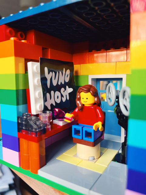 A photo showing a LEGO minifig with long brown hair sitting at a desk in a rainbow-colored home. On the desk you can see a “hard drive”, a cup of coffee and a keyboard. Behind the minifig there’s a projector and on the monitor there is a black and white sticker that reads “YUNO HOST” 