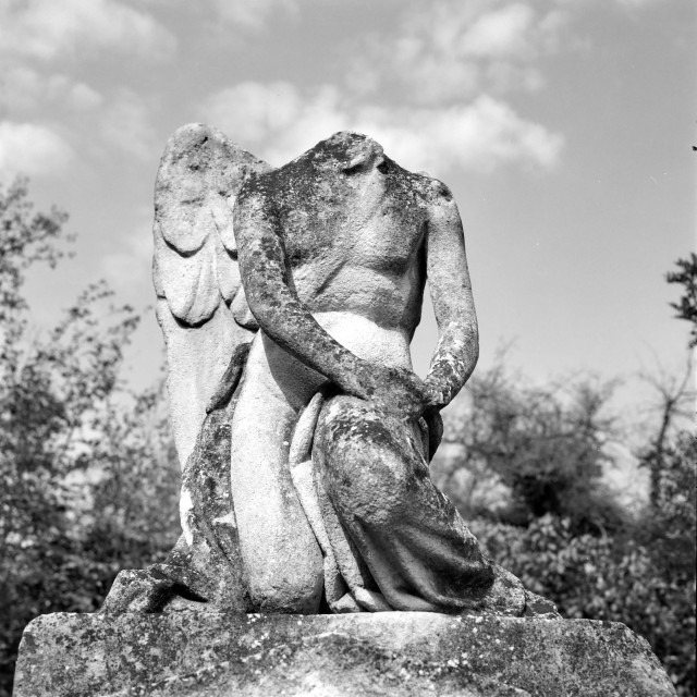 The black and white image shows a weathered, headless stone statue of a kneeling angel. One of the figure’s wings is missing, and its arms rest gently on its lap. The surface of the statue is rough and mottled with age, and the background reveals trees and a partly cloudy sky.
