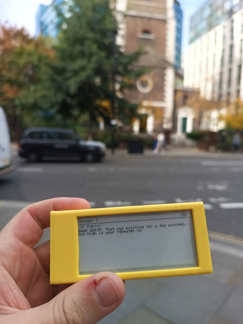Heltec client running MeshCore held up against a blurred London city streets background showing a message received