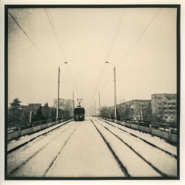 This lith print, captured with a Holga 120N, depicts a solitary tram moving toward the viewer along snow-covered tracks that stretch into the distance. The scene is enveloped in a pale, wintry haze, where the outlines of apartment blocks and lampposts fade into the mist. The soft focus and vignetting characteristic of the Holga lend the image a dreamlike, almost nostalgic atmosphere.

The grainy texture and warm-toned contrast of the lith process enhance the sense of melancholy and timelessness — as if the tram glides through a memory rather than a real city. The symmetrical composition and vanishing lines of the rails guide the eye toward the small, dark figure of the tram, creating a feeling of isolation and quiet endurance within an empty urban landscape.

It’s an image that feels both ghostly and poetic, blending the imperfection of toy-camera optics with the expressive tonal depth of lith printing — a quiet meditation on movement, solitude, and the passage of time.