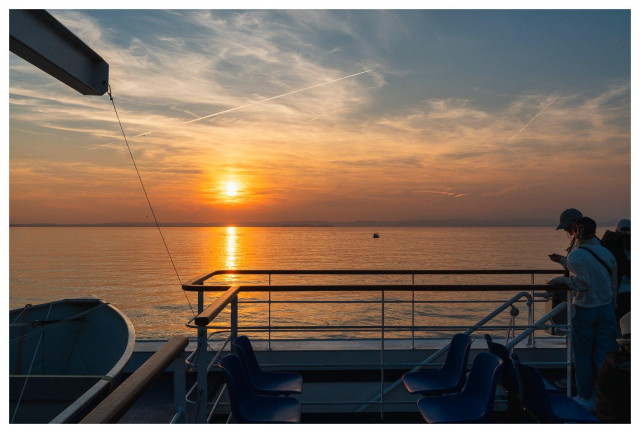 Bild in den Sonnenuntergang mit Aufbauten des Schiffs und Passagieren im Vordergrund.
