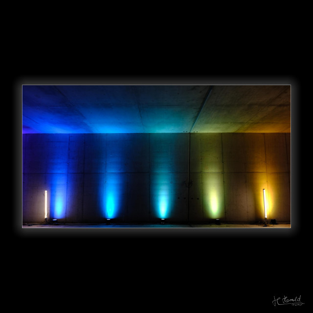Landscape image in a wide, square, black frame. View of a concrete wall and ceiling illuminated in colour by five spotlights. The colours change from blue to yellow from left to right. There is a vertical neon tube at each edge of the image. 