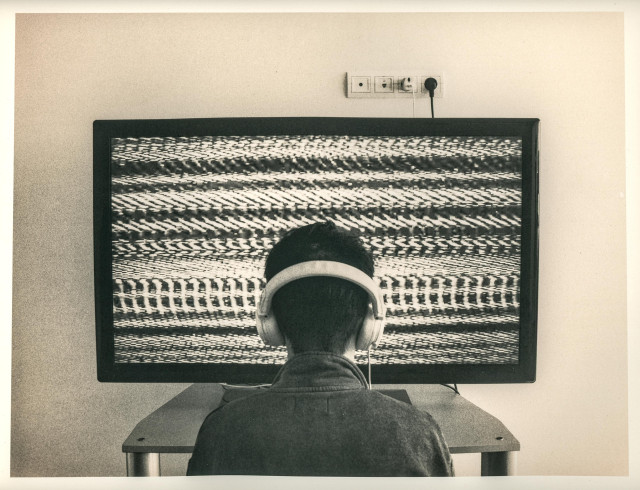 This lith print presents a strikingly minimal and symbolic scene.
At the center, a person with headphones sits facing a television screen that fills much of the frame. The TV displays a dense, rhythmic field of static interference — abstract, hypnotic waves of black and white noise. The viewer’s back is turned to us, emphasizing anonymity and detachment. The clean, clinical wall behind, with a single electrical socket and cord, heightens the sterile, almost laboratory-like atmosphere.

The image speaks of disconnection within connection — a modern ritual of isolation through technology. The lith process enhances this mood: the rough grain, high contrast, and muted tonal palette lend a sense of decay and psychological tension, as if this is a relic of an age that drowned in its own signals.