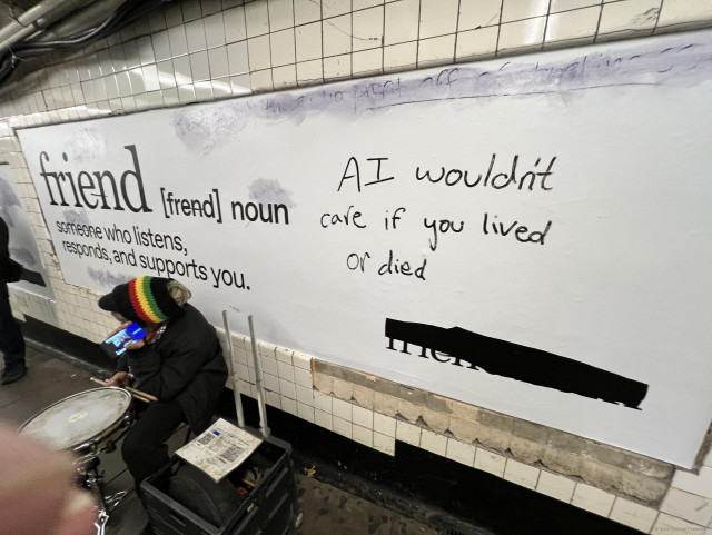 Photograph by Fenichel

In a NYC subway, on a platform, a man is seated low to the ground with a drum on a stand before him, stick in hand, wearing a dark coat and colorful wool hat.

Behind him is a long mural/advertisement (left half) and a hand-written comment/graffiti on the right side.

Advertisement, in the style of a dictionary definition, the biggest word being "friend"
 <<
[frend] noun
someone who listens,
responds and supports you.
>>

Graffiti/response/commentary:
"AI wouldn't care if you lived or died"

