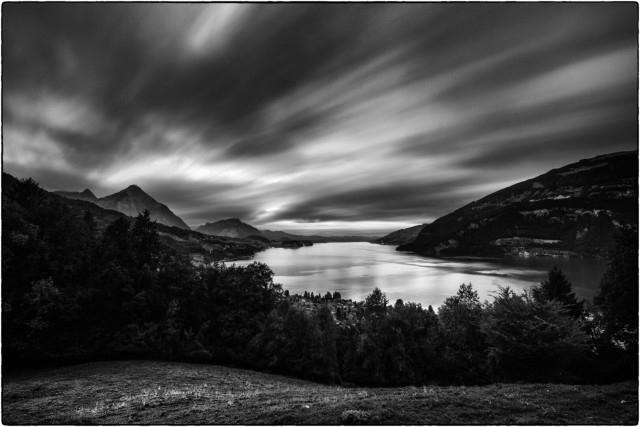 Ein weiter, ruhiger See liegt in einer Voralpenlandschaft, umgeben von dichten Wäldern und bewaldeten Bergen. Die Wasserfläche spiegelt das letzte Licht des Tages wider und schafft eine fast mystische Stimmung.

Am Himmel ziehen sich dynamische Wolkenformationen hin, die durch die Langzeitbelichtung weich und fliessend wirken. Die Bergsilhouetten im Hintergrund heben sich dunkel gegen den Himmel ab, während die Baumkronen im Vordergrund für Tiefe und Struktur sorgen.