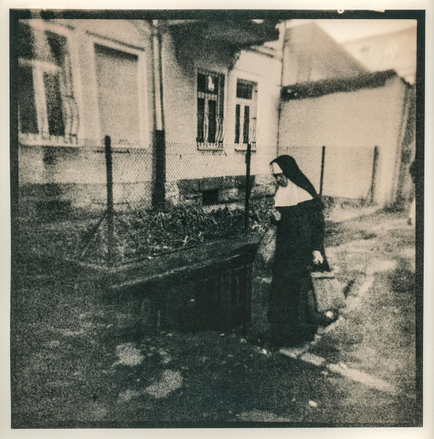 This lith print captures a fleeting street moment in Lviv — a solitary nun walking past an old courtyard enclosed by a wire fence. The scene is rendered in the characteristic lithographic palette of deep blacks and warm, grainy midtones, creating a sense of age and quiet melancholy. The soft focus and rough texture lend the image a dreamlike, almost ghostly atmosphere, as if drawn from a distant memory or a faded prayer.

The composition balances human presence and architectural silence: the nun’s dark habit contrasts with the pale, timeworn walls, while the surrounding emptiness enhances the feeling of solitude. The print’s gritty tonal structure — with its mottled shadows and veiled highlights — evokes both the spiritual austerity and poetic decay of old Lviv.
