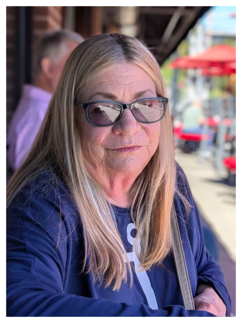 A woman with long blonde hair and sunglasses stands sits outside a cafe at an unseen table, wearing a navy blue shirt with a white anchor design. She has a neutral expression. The background shows blurred people, red umbrellas, and outdoor seating on a sunny day.