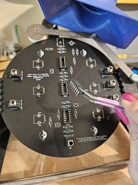 Black circular PCB, mid build. PCB is gripped in a holder frame. My hand is partially visible, holding a 14 pin chip between the grips of a pink pair of tweezers. Hot pink nail polish. The opening of a blue fume extractor can be seen in the top right.