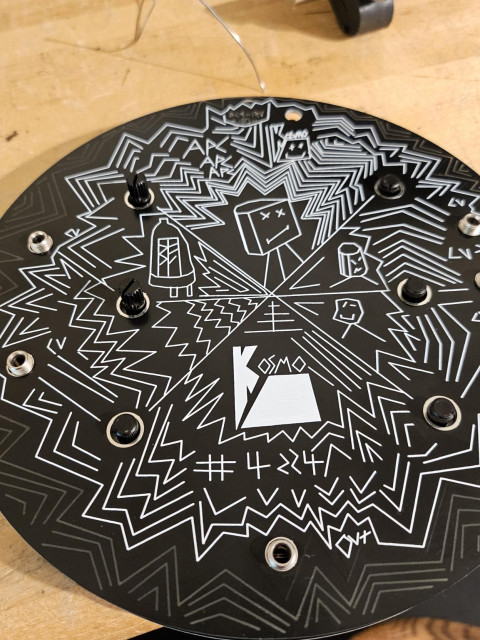 Top side of the synthesizer with potentiometers poking through a circular black panel of white, graphic lines making up a design of visually busy surface art.